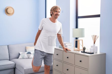 Téléchargez les photos : Jeune homme blond souriant confiant étirement jambe à la maison - en image libre de droit
