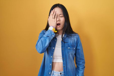 Foto de Mujer asiática joven de pie sobre fondo amarillo bostezando cansado cubriendo media cara, ojo y boca con la mano. cara duele en el dolor. - Imagen libre de derechos