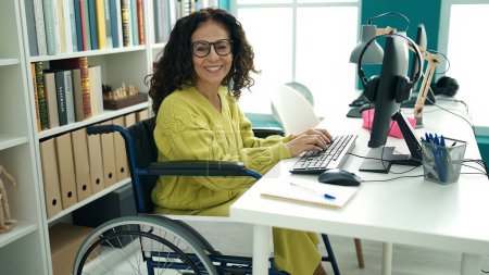 Foto de Middle age hispanic woman teacher using computer sitting on wheelchair at library university - Imagen libre de derechos