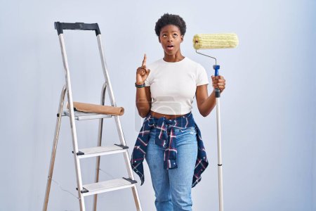 Téléchargez les photos : Femme afro-américaine tenant peintre rouleau étonné et surpris regardant vers le haut et pointant avec les doigts et les bras levés. - en image libre de droit