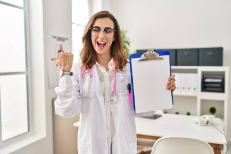 Photo for Young doctor woman holding clipboard pointing thumb up to the side smiling happy with open mouth - Royalty Free Image