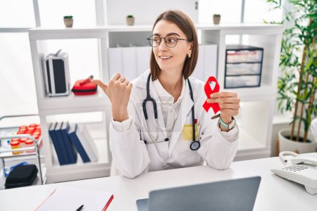 Photo for Young caucasian doctor woman holding support red ribbon pointing thumb up to the side smiling happy with open mouth - Royalty Free Image