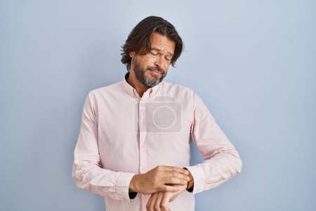 Foto de Hombre guapo de mediana edad que usa un fondo de camisa elegante comprobando la hora en el reloj de pulsera, relajado y seguro - Imagen libre de derechos