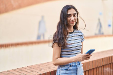 Téléchargez les photos : Jeune fille hispanique souriant confiant en utilisant smartphone à la rue - en image libre de droit