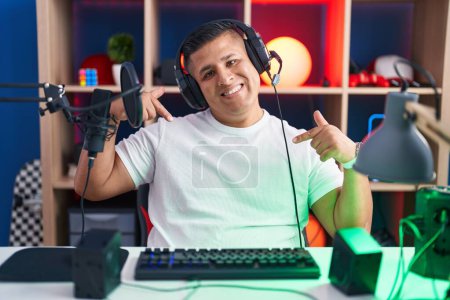 Foto de Joven hispano jugando videojuegos con cara sonriente, señalándose con los dedos orgullosos y felices. - Imagen libre de derechos