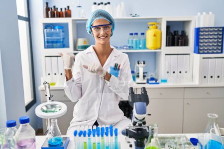 Foto de Mujer morena trabajando en el laboratorio científico apuntando hacia atrás con la mano y los pulgares hacia arriba, sonriendo confiada - Imagen libre de derechos