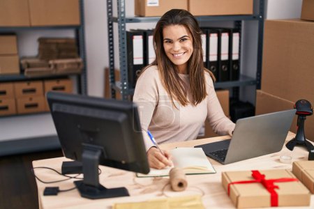 Photo for Young beautiful hispanic woman ecommerce business worker using laptop writing on notebook at office - Royalty Free Image