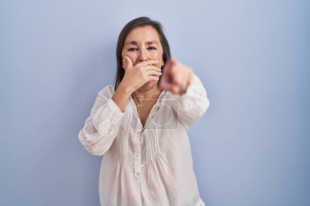 Photo for Middle age hispanic woman standing over blue background laughing at you, pointing finger to the camera with hand over mouth, shame expression - Royalty Free Image