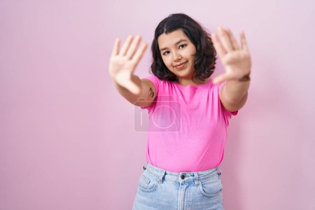 Foto de Mujer hispana joven de pie sobre fondo rosa haciendo marco usando las manos palmas y los dedos, perspectiva de la cámara - Imagen libre de derechos