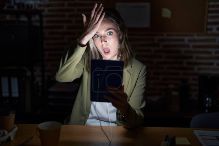 Photo for Blonde caucasian woman working at the office at night surprised with hand on head for mistake, remember error. forgot, bad memory concept. - Royalty Free Image
