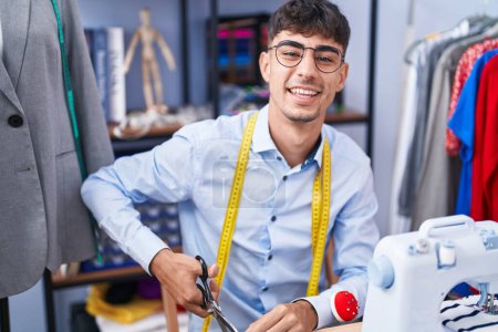 Téléchargez les photos : Jeune homme hispanique tailleur souriant tissu de coupe confiant à la boutique de tailleur - en image libre de droit