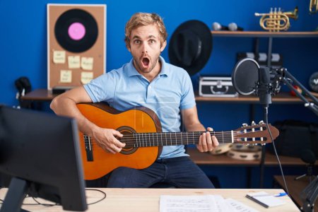 Téléchargez les photos : Homme caucasien jouant de la guitare classique au studio de musique effrayé et choqué par la surprise et l'expression étonnante, la peur et le visage excité. - en image libre de droit