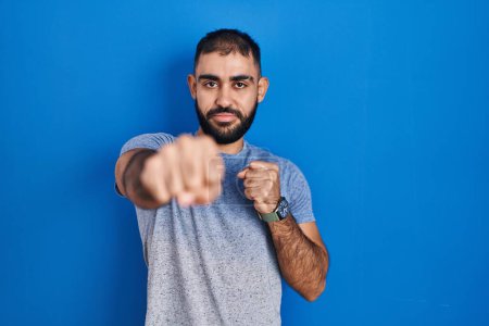Photo for Middle east man with beard standing over blue background punching fist to fight, aggressive and angry attack, threat and violence - Royalty Free Image
