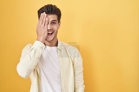 Foto de Young hispanic man standing over yellow background covering one eye with hand, confident smile on face and surprise emotion. - Imagen libre de derechos