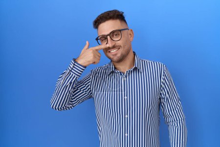 Téléchargez les photos : Homme hispanique avec la barbe portant des lunettes pointant avec le doigt de la main vers le visage et le nez, souriant gai. concept de beauté - en image libre de droit