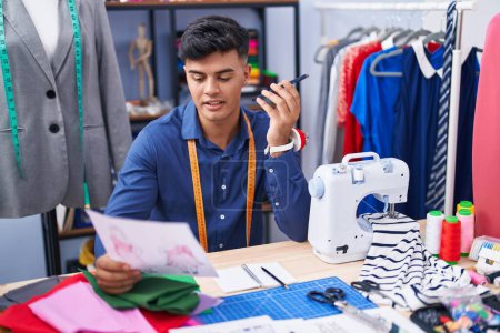 Téléchargez les photos : Jeune homme hispanique tailleur parler sur smartphone regarder conception de vêtements à l'usine de vêtements - en image libre de droit