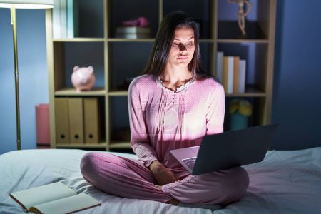 Photo for Young hispanic woman using computer laptop on the bed relaxed with serious expression on face. simple and natural looking at the camera. - Royalty Free Image