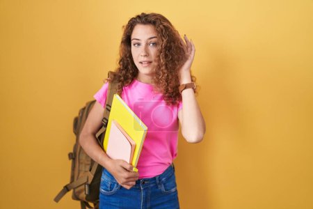 Photo for Young caucasian woman wearing student backpack and holding books smiling with hand over ear listening an hearing to rumor or gossip. deafness concept. - Royalty Free Image