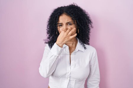 Foto de Hispanic woman with curly hair standing over pink background smelling something stinky and disgusting, intolerable smell, holding breath with fingers on nose. bad smell - Imagen libre de derechos