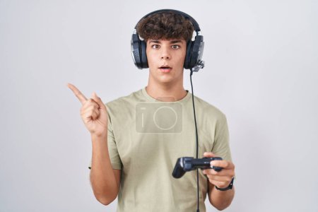Téléchargez les photos : Adolescent hispanique jouant contrôleur de tenue de jeu vidéo surpris pointant du doigt sur le côté, bouche ouverte expression étonnée. - en image libre de droit