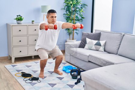 Photo for Young latin man training leg exercise using dumbbells at home - Royalty Free Image
