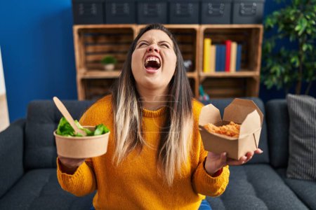 Photo for Young hispanic woman holding healthy salad and fried chicken wings angry and mad screaming frustrated and furious, shouting with anger looking up. - Royalty Free Image