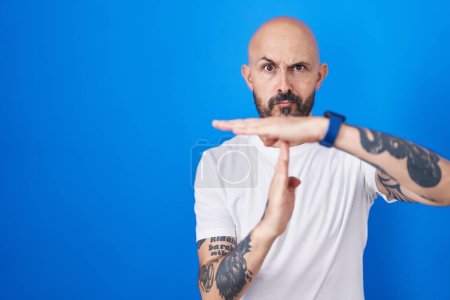 Foto de Hombre hispano con tatuajes de pie sobre fondo azul haciendo un gesto de tiempo fuera con las manos, rostro frustrado y serio - Imagen libre de derechos