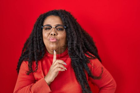 Téléchargez les photos : Plus taille hispanique femme debout sur fond rouge pensée concentrée sur le doute avec le doigt sur le menton et la recherche jusqu'à se demander - en image libre de droit
