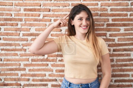 Photo for Young brunette woman standing over bricks wall smiling pointing to head with one finger, great idea or thought, good memory - Royalty Free Image