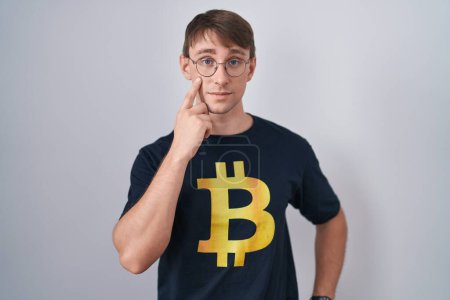Foto de Hombre rubio caucásico con camiseta bitcoin apuntando al ojo observándote gesto, expresión sospechosa - Imagen libre de derechos