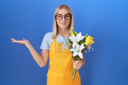 Photo for Young caucasian woman wearing florist apron holding flowers smiling cheerful presenting and pointing with palm of hand looking at the camera. - Royalty Free Image