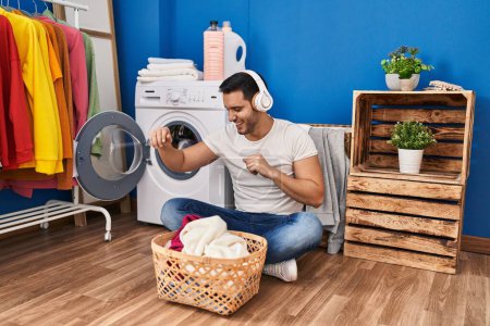 Foto de Young hispanic man dancing and listening to music washing clothes at laundry room - Imagen libre de derechos