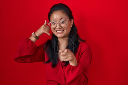 Téléchargez les photos : Asiatique jeune femme debout sur fond rouge souriant faire parler au téléphone geste et vous pointant du doigt. Appelle-moi. - en image libre de droit