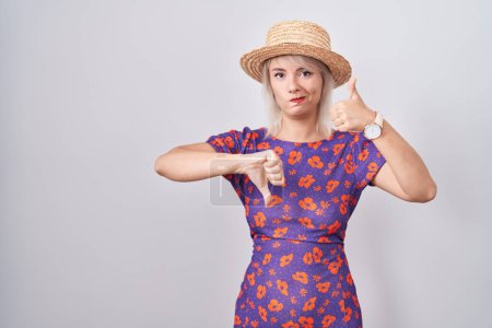 Foto de Mujer caucásica joven con vestido de flores y sombrero de verano haciendo pulgares arriba y abajo, desacuerdo y expresión de acuerdo. conflicto loco - Imagen libre de derechos