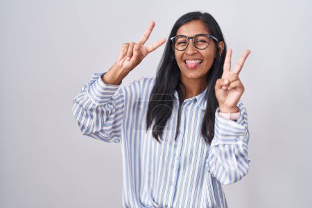 Téléchargez les photos : Jeune femme hispanique portant des lunettes souriant avec la langue montrant les doigts des deux mains faisant signe de victoire. numéro deux. - en image libre de droit