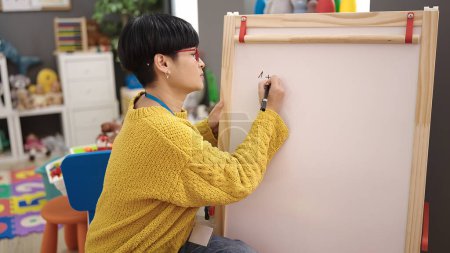 Photo for Young chinese woman preschool teacher sitting on chair writing on chalkboard at kindergarten - Royalty Free Image