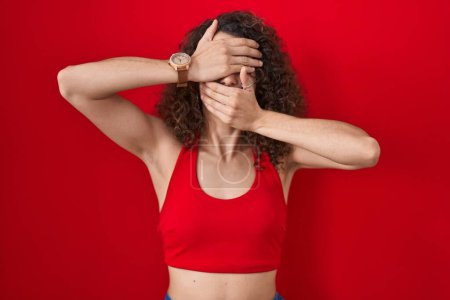 Photo for Hispanic woman with curly hair standing over red background covering eyes and mouth with hands, surprised and shocked. hiding emotion - Royalty Free Image