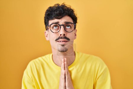 Téléchargez les photos : Homme hispanique portant des lunettes debout sur fond jaune mendier et prier avec les mains ainsi que l'expression de l'espoir sur le visage très émotionnel et inquiet. mendier. - en image libre de droit