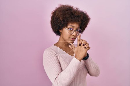 Foto de Joven mujer afroamericana de pie sobre fondo rosa sosteniendo un arma simbólica con gesto de mano, jugando a matar armas de fuego, cara enojada - Imagen libre de derechos