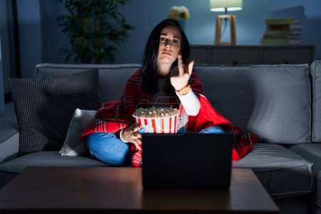 Photo for Hispanic woman eating popcorn watching a movie on the sofa showing middle finger, impolite and rude fuck off expression - Royalty Free Image
