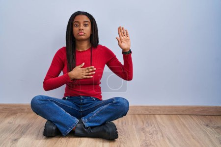 Téléchargez les photos : Jeune afro-américain avec des tresses assises sur le sol à la maison jurant avec la main sur la poitrine et la paume ouverte, faisant un serment de fidélité - en image libre de droit