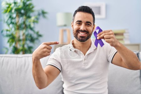 Téléchargez les photos : Jeune homme hispanique avec barbe tenant un ruban violet conscience pointant du doigt un sourire heureux et fier - en image libre de droit
