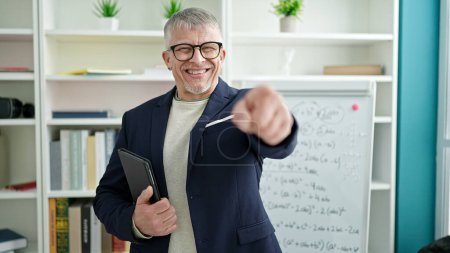 Téléchargez les photos : Moyen âge homme aux cheveux gris enseignant tenant pavé tactile pointant du doigt à la caméra à l'université salle de classe - en image libre de droit
