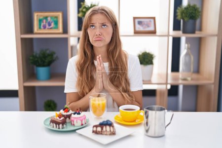 Photo for Young caucasian woman eating pastries t for breakfast begging and praying with hands together with hope expression on face very emotional and worried. begging. - Royalty Free Image