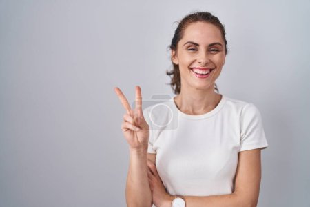 Téléchargez les photos : Belle femme brune debout sur un arrière-plan isolé souriant avec un visage heureux clin d'oeil à la caméra faisant signe de victoire avec les doigts. numéro deux. - en image libre de droit
