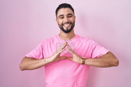 Foto de Joven hispano de pie sobre el fondo rosa manos juntas y los dedos cruzados sonriendo relajado y alegre. éxito y optimismo - Imagen libre de derechos