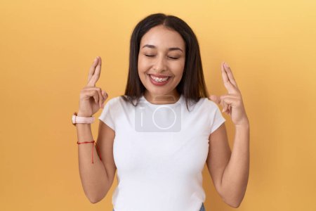 Foto de Mujer árabe joven vistiendo camiseta blanca casual sobre fondo amarillo gesto dedo cruzado sonriendo con esperanza y los ojos cerrados. suerte y concepto supersticioso. - Imagen libre de derechos