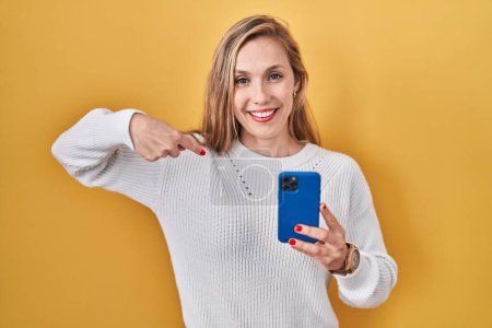 Téléchargez les photos : Jeune femme blonde utilisant un smartphone dactylographier message regardant confiant avec le sourire sur le visage, se montrant du doigt fier et heureux. - en image libre de droit