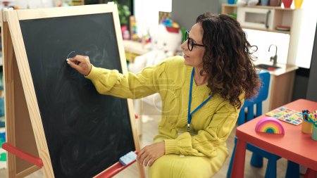 Photo for Middle age hispanic woman preschool teacher writing on blackboard at kindergarten - Royalty Free Image