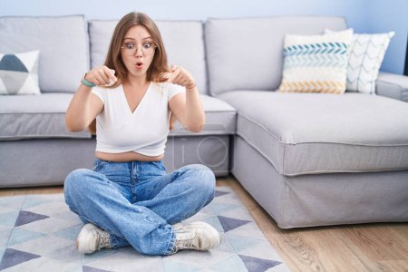 Photo for Young caucasian woman sitting on the floor at the living room pointing down with fingers showing advertisement, surprised face and open mouth - Royalty Free Image